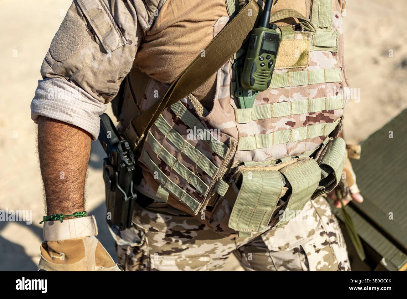 Primo piano di un uomo che indossa un giubbotto tattico in un ambiente desertico. L'attrezzatura mimetica è dotata di una radio che mette in evidenza la prontezza e la comunicazione Foto Stock