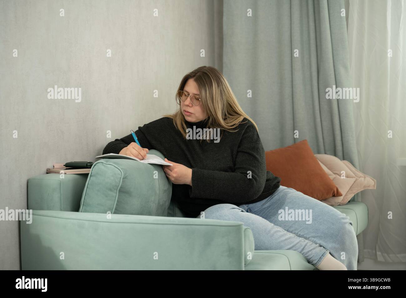 Una donna con gli occhiali scrive in un notebook mentre è seduta comodamente su un divano color pastello a casa. Con un comodo pullover e un paio di jeans, casual Foto Stock