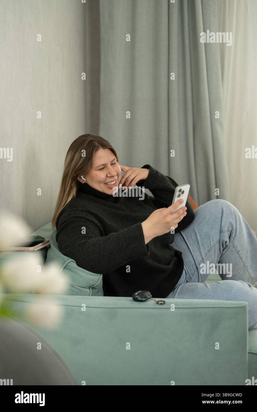 Una donna allegra siede comodamente su un divano color pastello, utilizzando il suo smartphone e sorridendo. Vestita in modo informale con un maglione nero e un paio di jeans, le piace Foto Stock