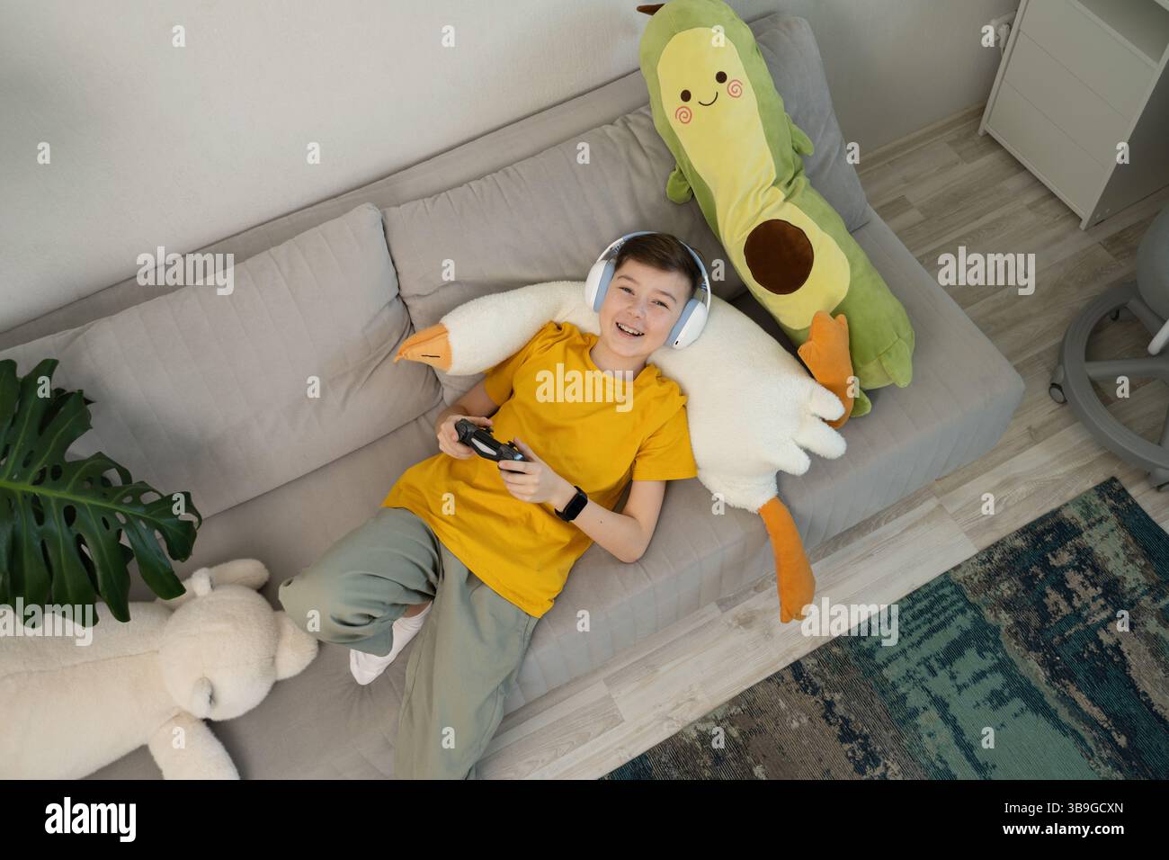 Un ragazzo felice che indossa le cuffie gioca ai videogiochi mentre si rilassa su un comodo divano. Circondato da peluche, ama una rilassante sessione di gioco a casa Foto Stock