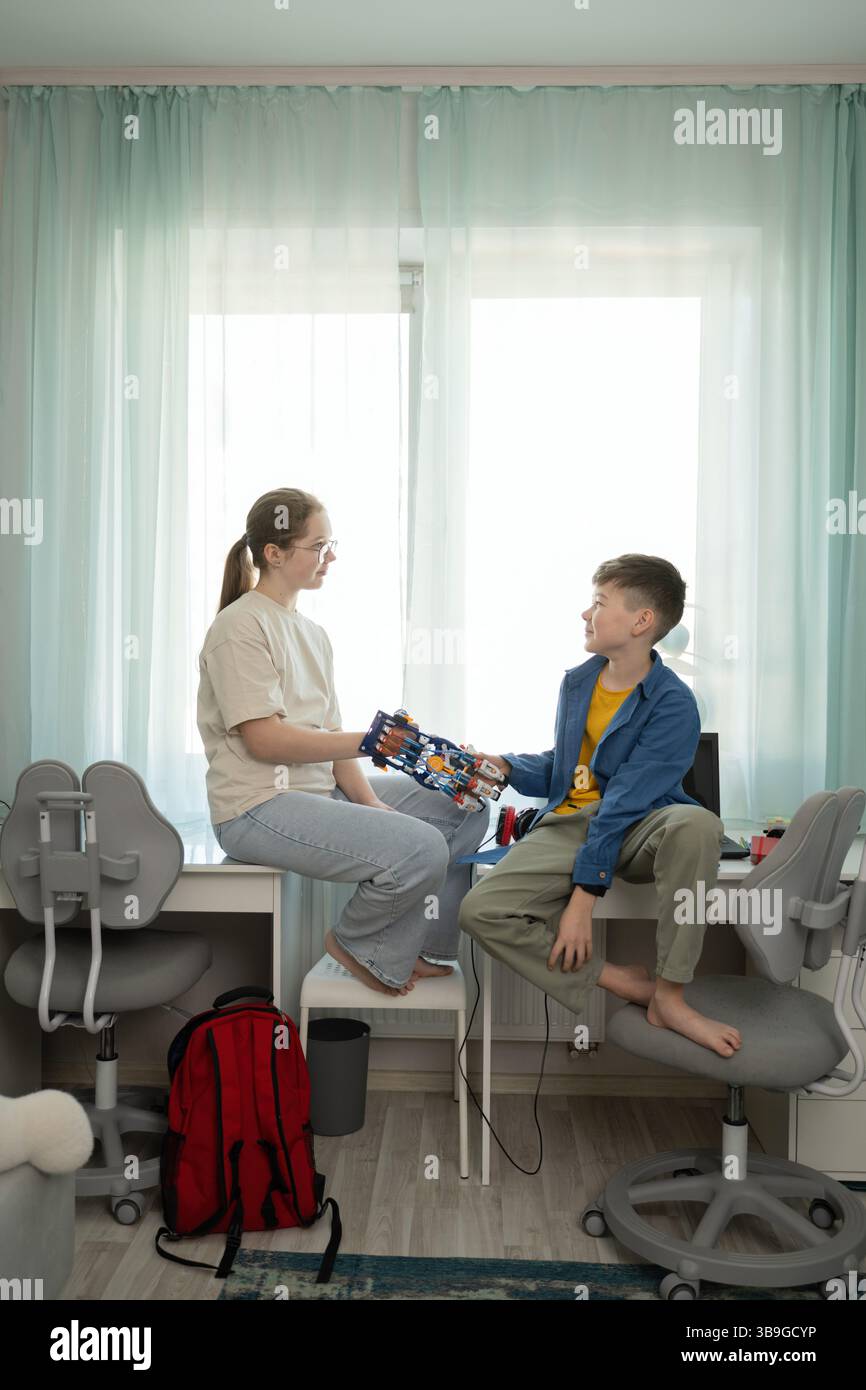 Un ragazzo e una ragazza condividono un momento con una mano robotica. L'ambiente e' una sala accogliente e luminosa piena di materiali educativi che simboleggiano la creatività e. Foto Stock