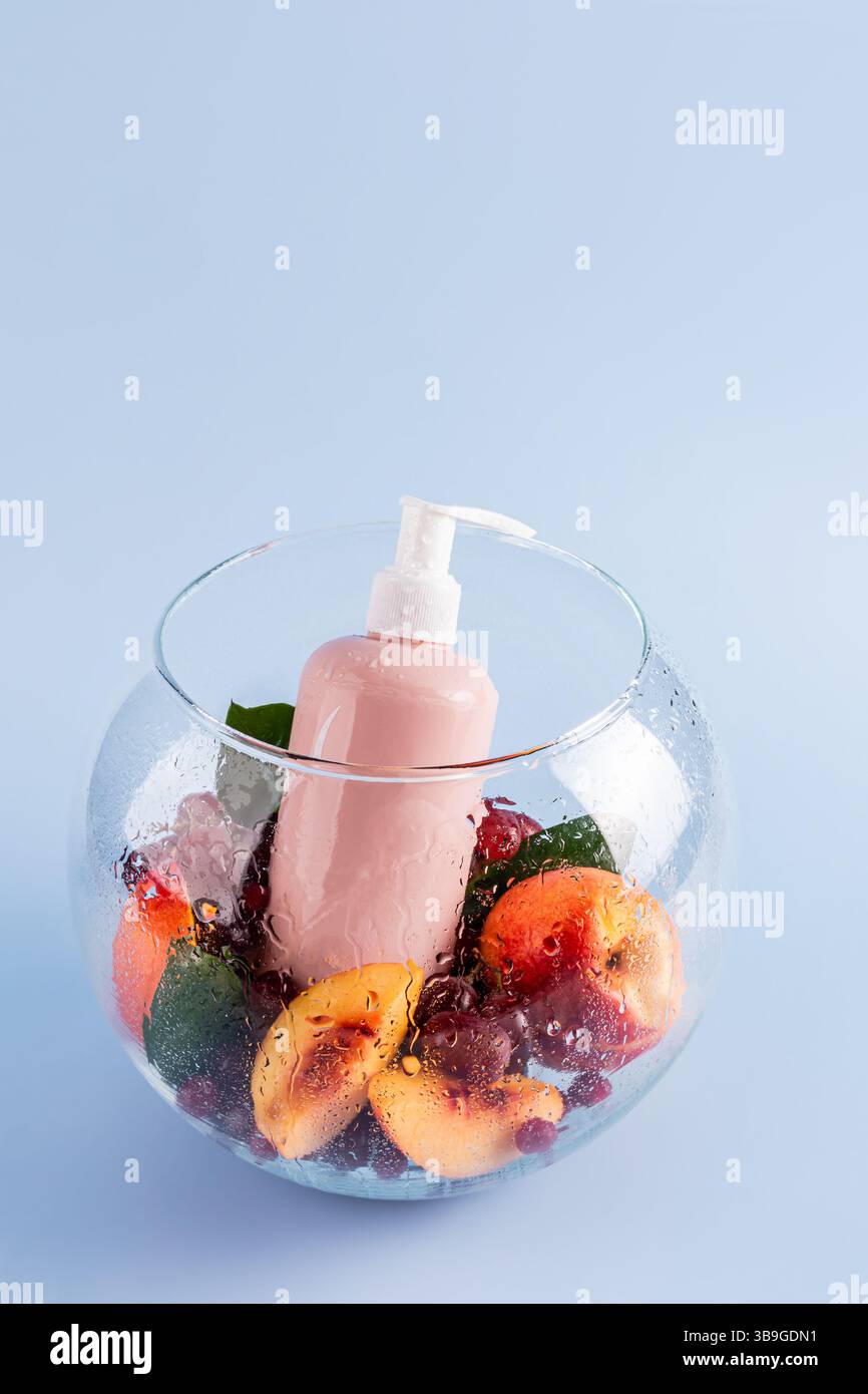 Una rinfrescante bottiglia di gel doccia rosa si trova in una ciotola di vetro circondata da pesche e frutti di bosco, creando un ambiente estetico e aromatico simile a una spa Foto Stock