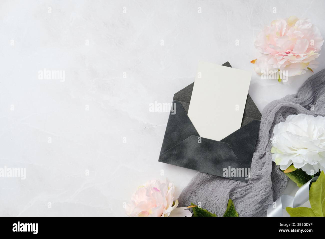 Elegante cartoleria per matrimoni con una busta di classe con un invito vuoto circondata da delicati fiori bianchi su una superficie in marmo morbido, ideale per chi ama Foto Stock