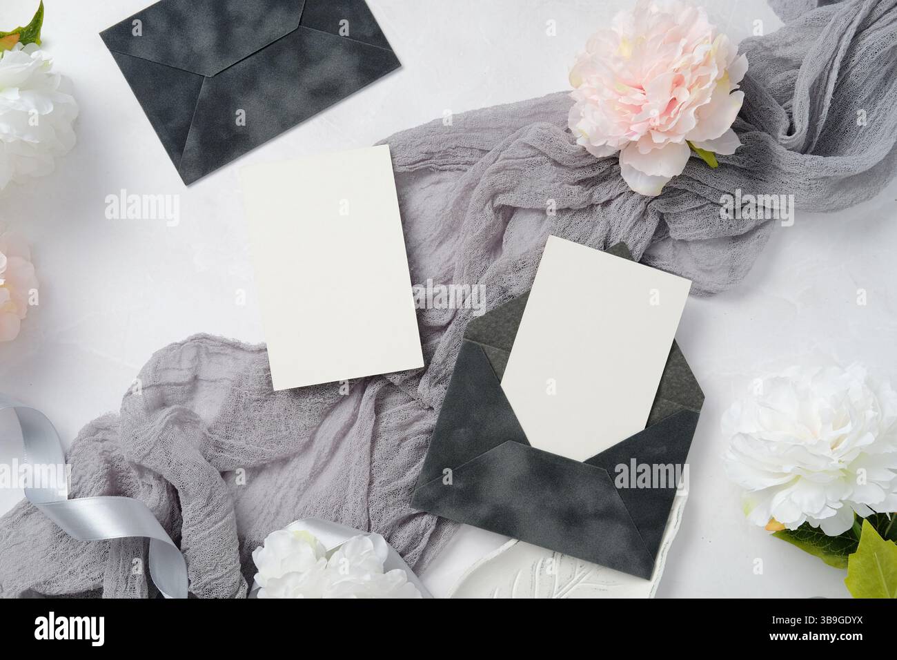 Elegante set di cancelleria per matrimoni con buste scure e carte bianche. Accompagnato da un morbido tessuto grigio e fiori di peonia bianca, crea un sofisticato Foto Stock