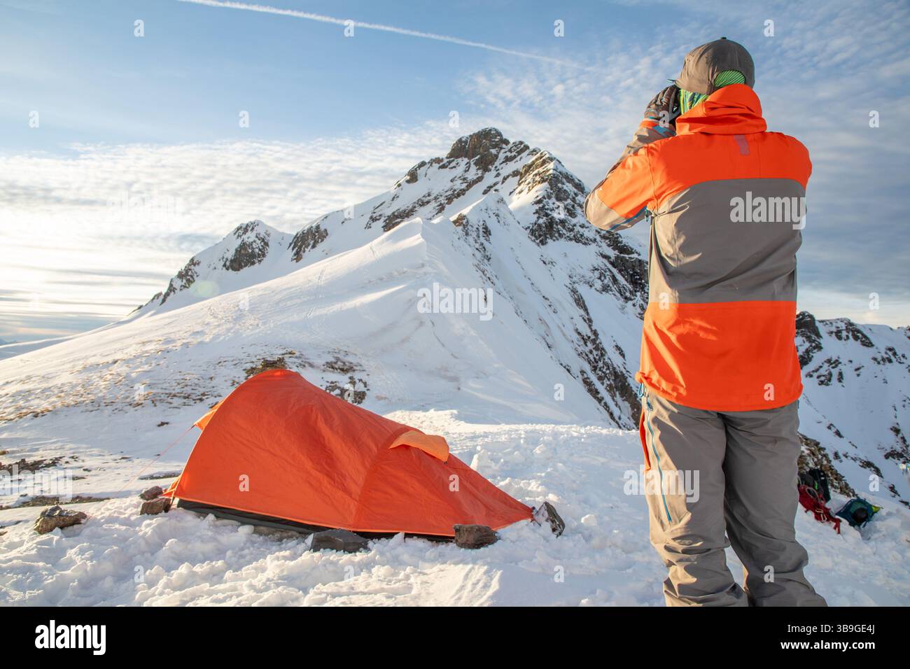 Una vivace scena di uno sciatore con un vestito arancione che esplora una montagna innevata. Una tenda arancione brillante contrasta con il paesaggio innevato sotto una B. Foto Stock