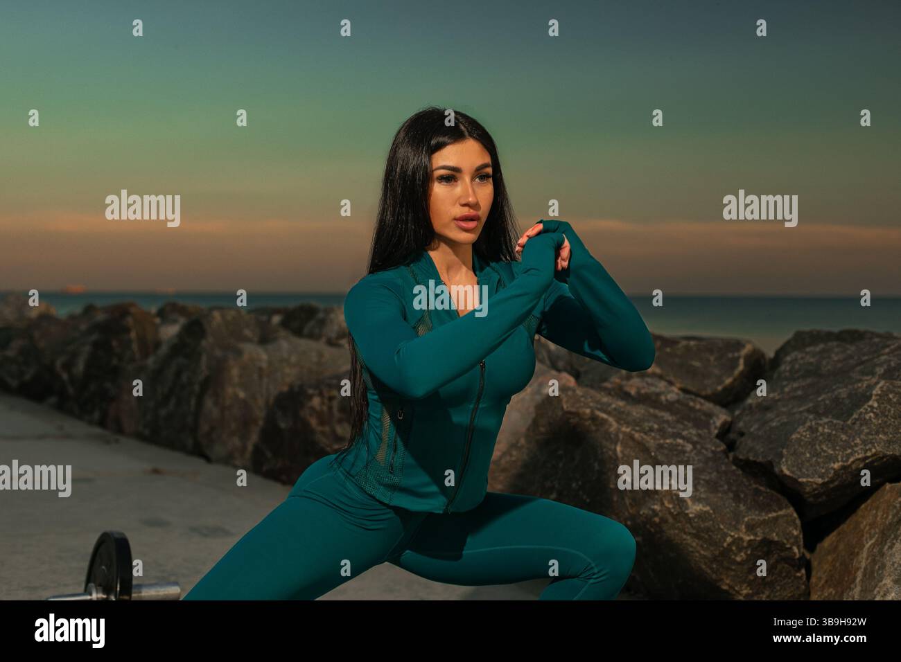 il modello fitness è impegnato in una posa dinamica di allenamento su un sentiero costiero circondato da rocce e oceano tranquillo. il tramonto crea una scena che arricchisce lo sfondo. Foto Stock