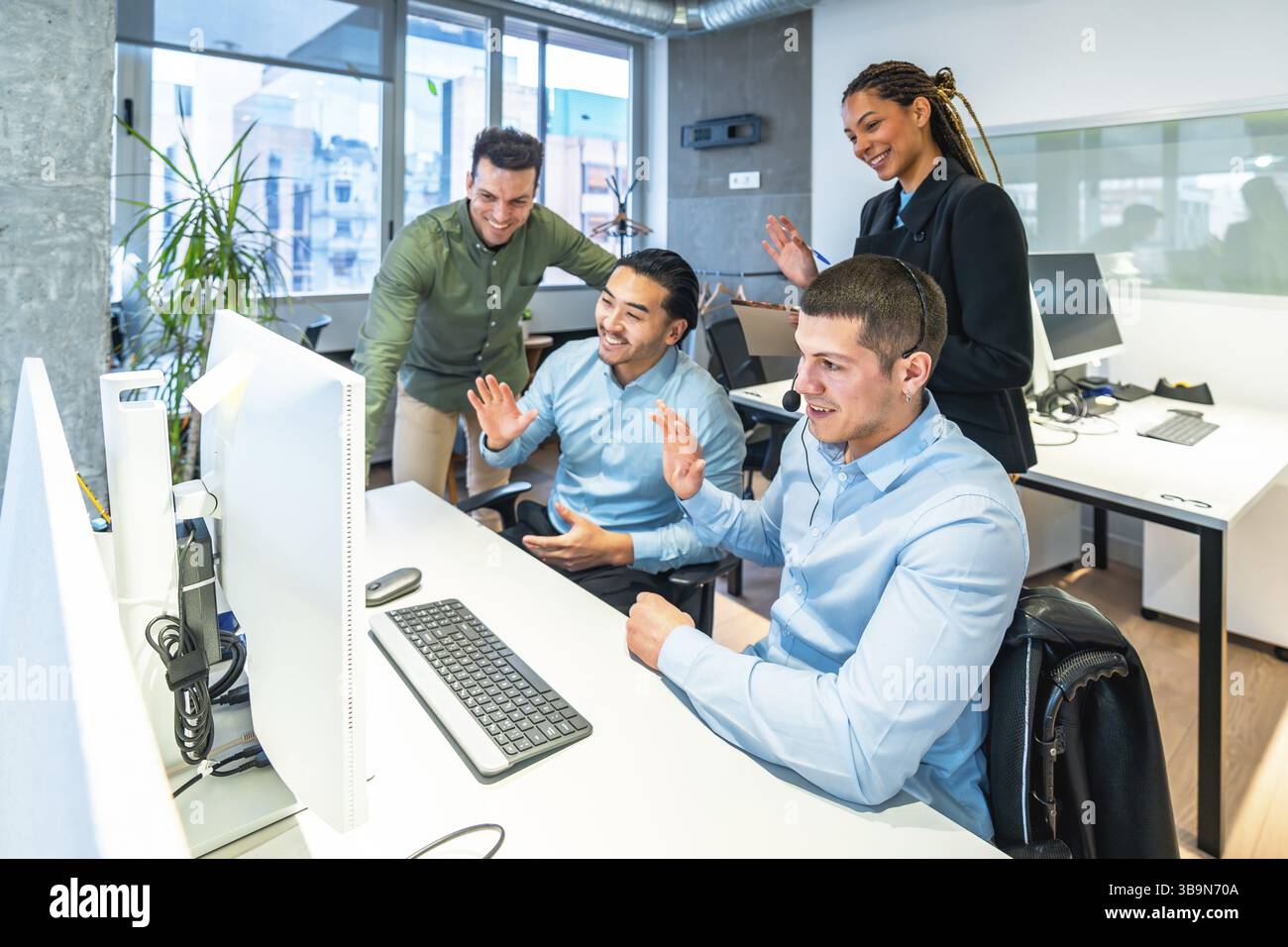 I colleghi d'affari salutano con entusiasmo il computer durante una videochiamata coinvolgente in un ambiente d'ufficio luminoso e moderno Foto Stock