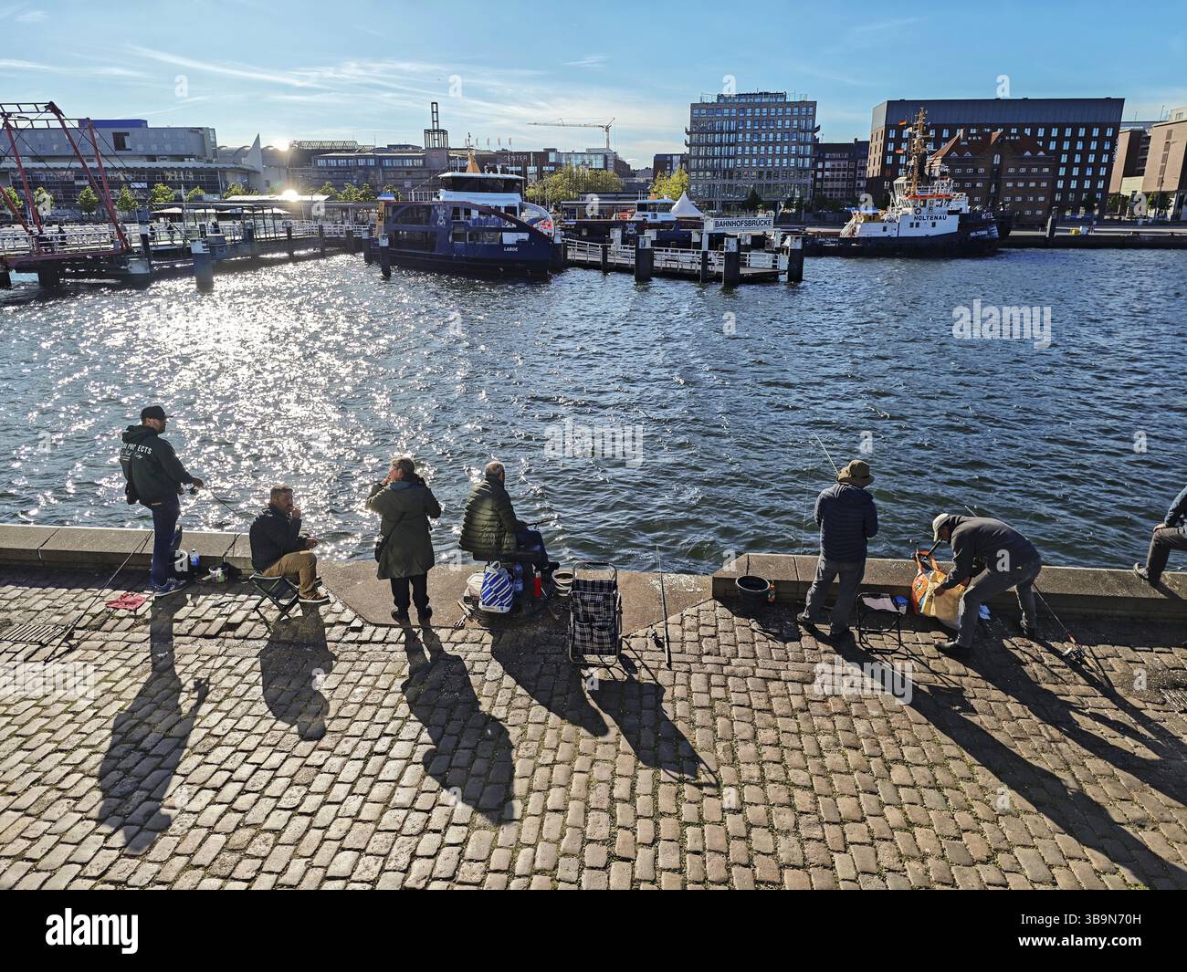 Porto di Kiel alla fine del fiordo di Kiel, vista dalla riva orientale con i pescatori alla riva occidentale con la stazione ferroviaria principale, Hoernbruecke B. Foto Stock