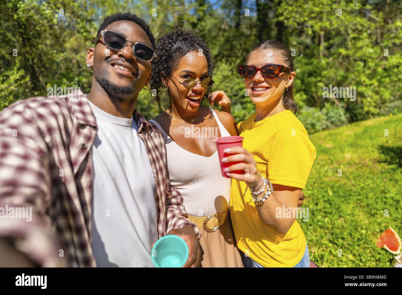 Tre amici allegri si godono un picnic, si scattano un selfie e tengono in mano tazze colorate in una giornata di sole nel parco Foto Stock