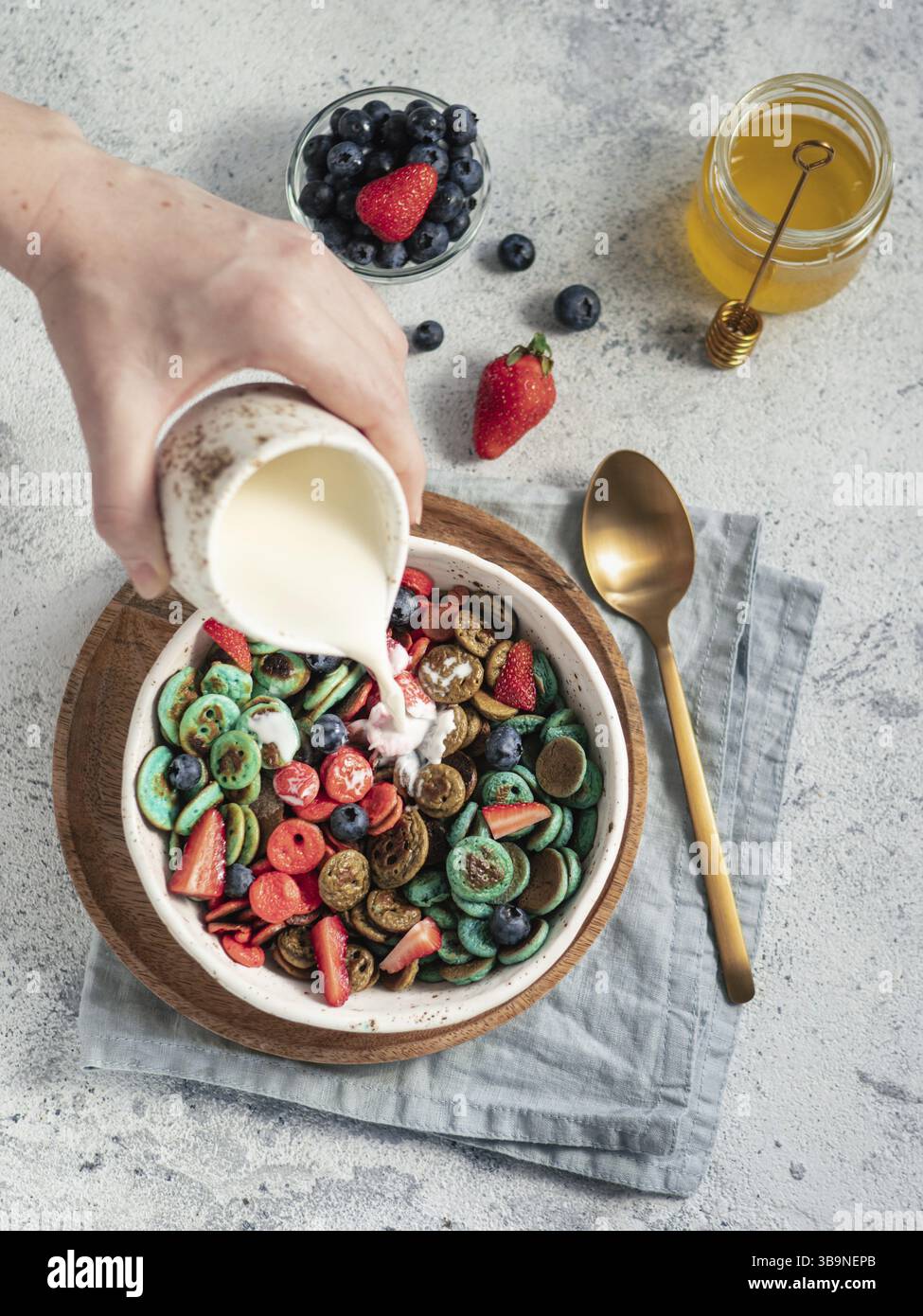 Cibo alla moda - pancake con cereali. Versare il latte su un mucchio di mini pancake di cereali multicolore in un recipiente. Minuscole frittelle di cereali con frutti di bosco in un piatto artigianale di oltre g Foto Stock