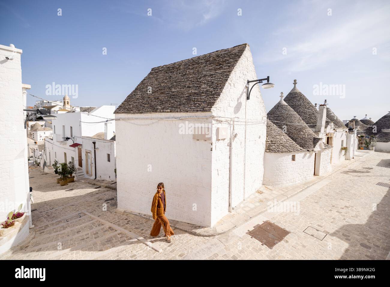 Donna che cammina lungo un'affascinante strada acciottolata ad Alberobello, circondata da iconiche case di trulli imbiancate con tetti conici sotto un cielo azzurro Foto Stock