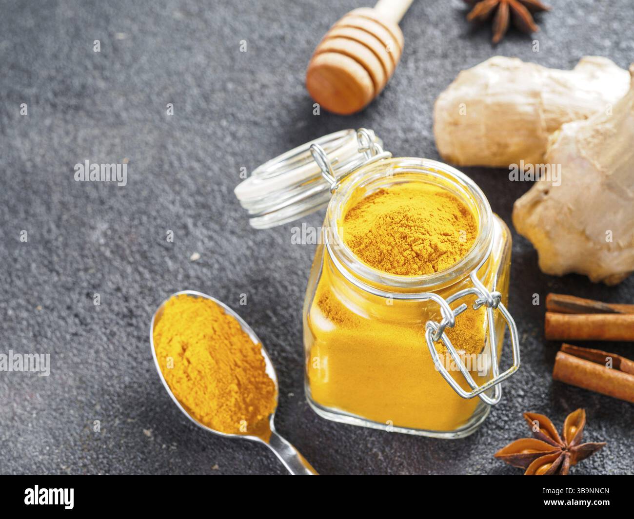Polvere di curcuma secca in vaso di vetro e cucchiaio su fondo di cemento nero. Ingredienti per latte dorato, latte di curcuma o bevanda detox - cucchiaino di miele, gin Foto Stock