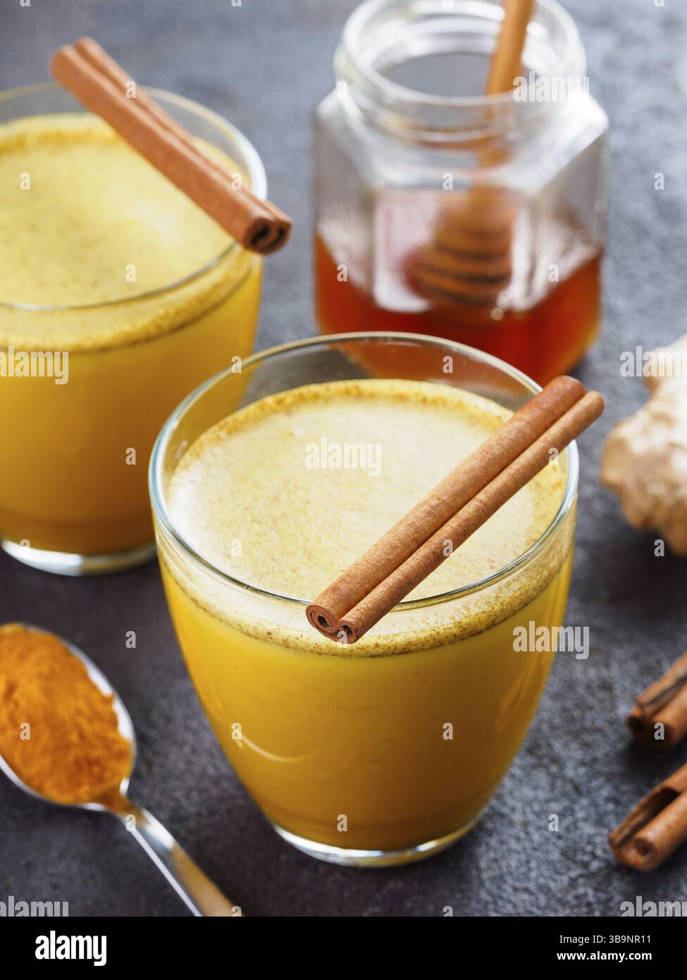 Latte di curcuma dorato in vetro da bere sano.latte dorato con curcuma, radice di zenzero, bastoncini di cannella, polvere di curcuma secca e miele su cemento nero Foto Stock