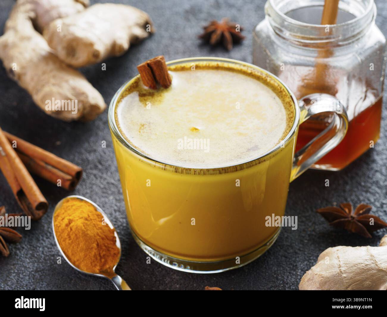 Latte di curcuma dorato in tazza di vetro. Latte dorato con curcuma, radice di zenzero, bastoncini di cannella, polvere di curcuma essiccata e miele su cem nero Foto Stock