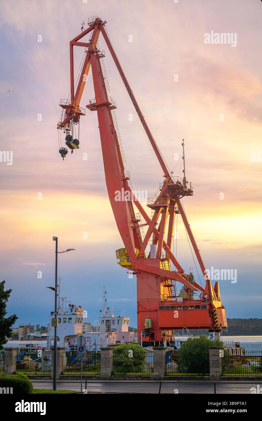 Una grande gru arancione è posizionata nel porto di Pola, sollevando attivamente il carico sotto un cielo vivace durante il tramonto. Foto Stock