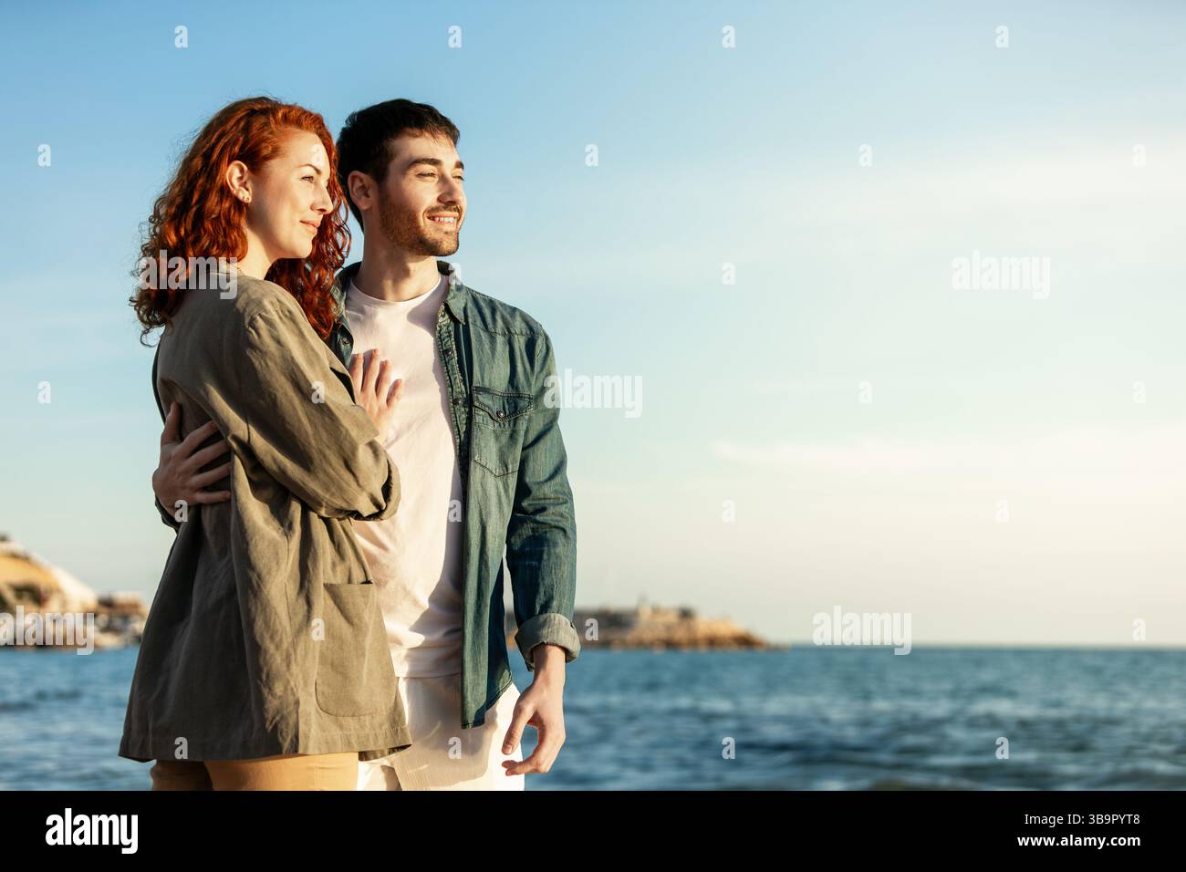 Una giovane coppia si trova in riva al mare sorridente e avvolgente mentre guarda in lontananza, godendosi un momento di sole tranquillo vicino all'acqua. LIF Foto Stock