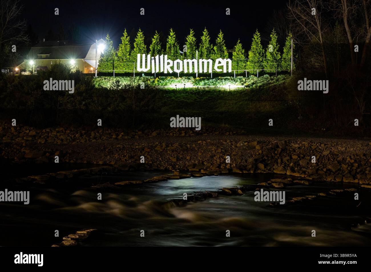 Foto del cartello di benvenuto vicino alle rapide del fiume a Frankenmuth, Michigan, illuminato di notte con acqua tranquilla e luce calda. Foto Stock