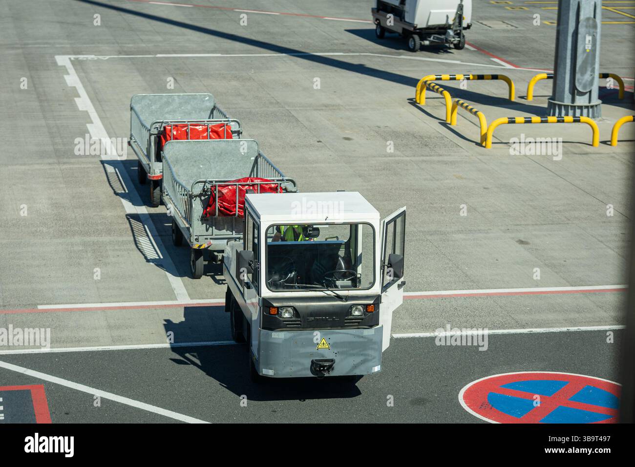 Un veicolo per il trasporto bagagli su un asfalto dell'aeroporto tira due carrelli con borse rosse, vicino a una zona "senza parcheggio". Le barriere gialle e un palo sono visibili, con una Foto Stock
