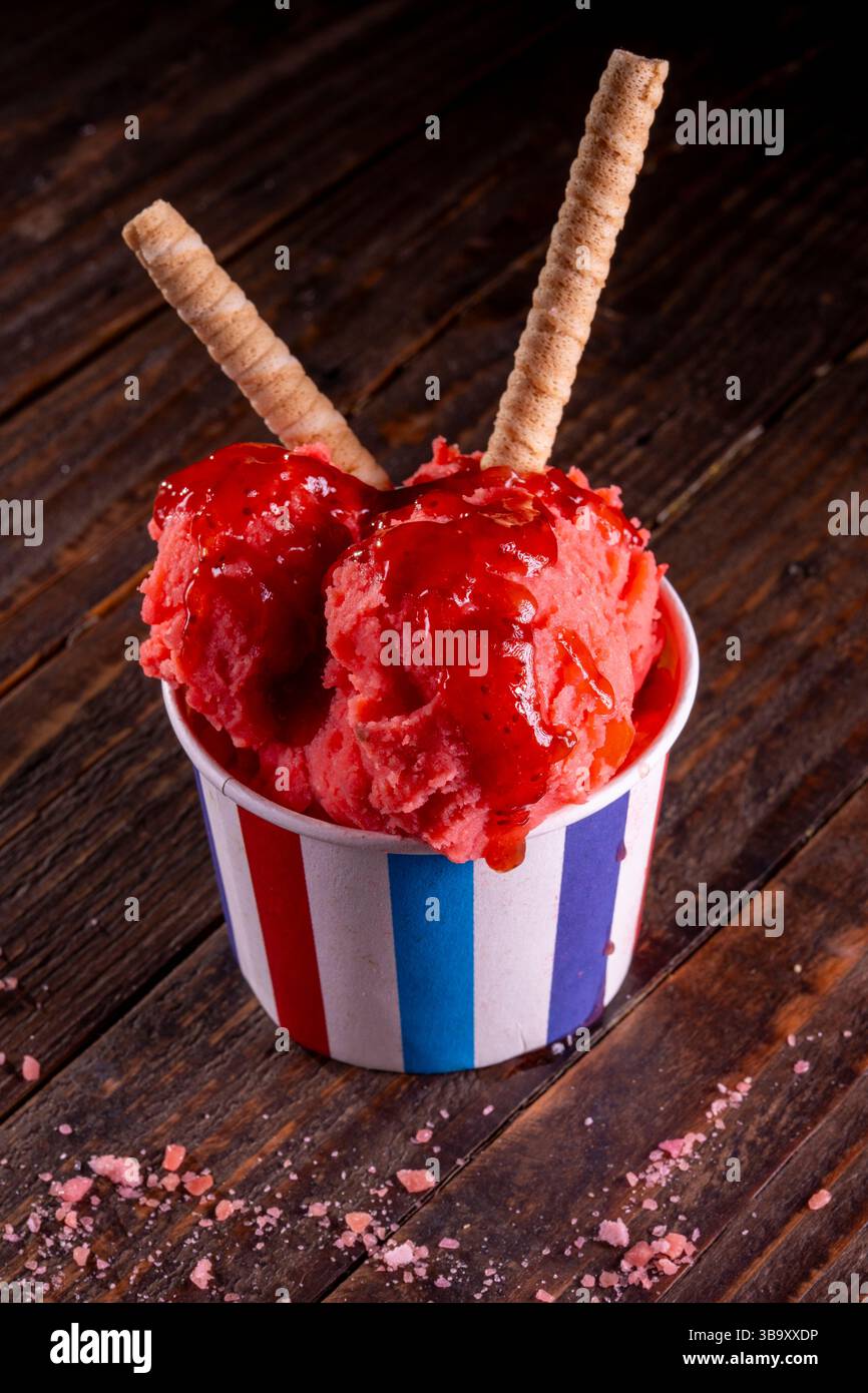 Primo piano di vibranti sorbetti alle fragole con marmellata servita in una tazza di carta colorata su un tavolo di legno scuro, dessert estivo Foto Stock