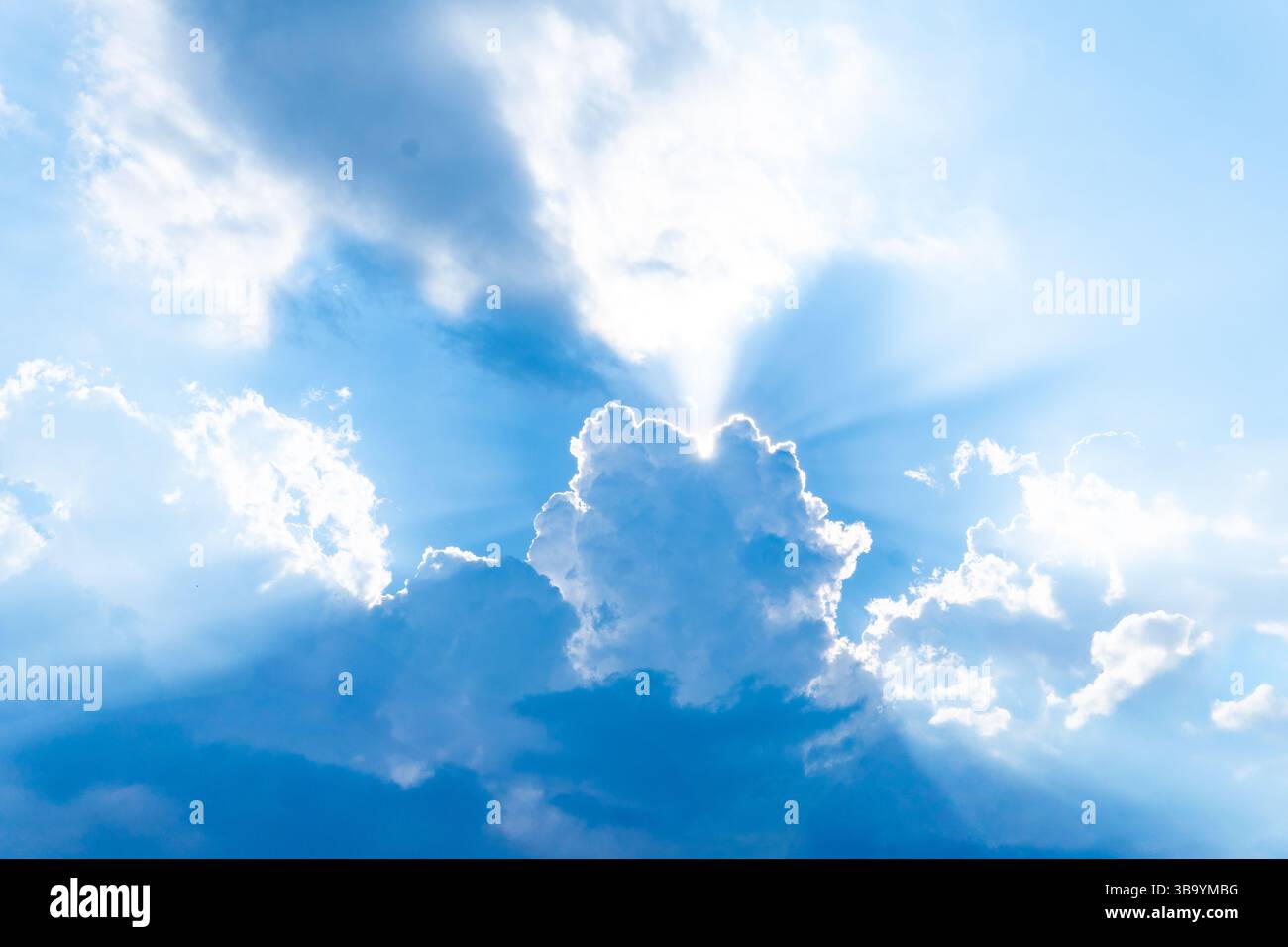 Un cielo blu vibrante e la luce del sole si diffondono da dietro una nuvola centrale e più grande, creando raggi luminosi. Foto Stock