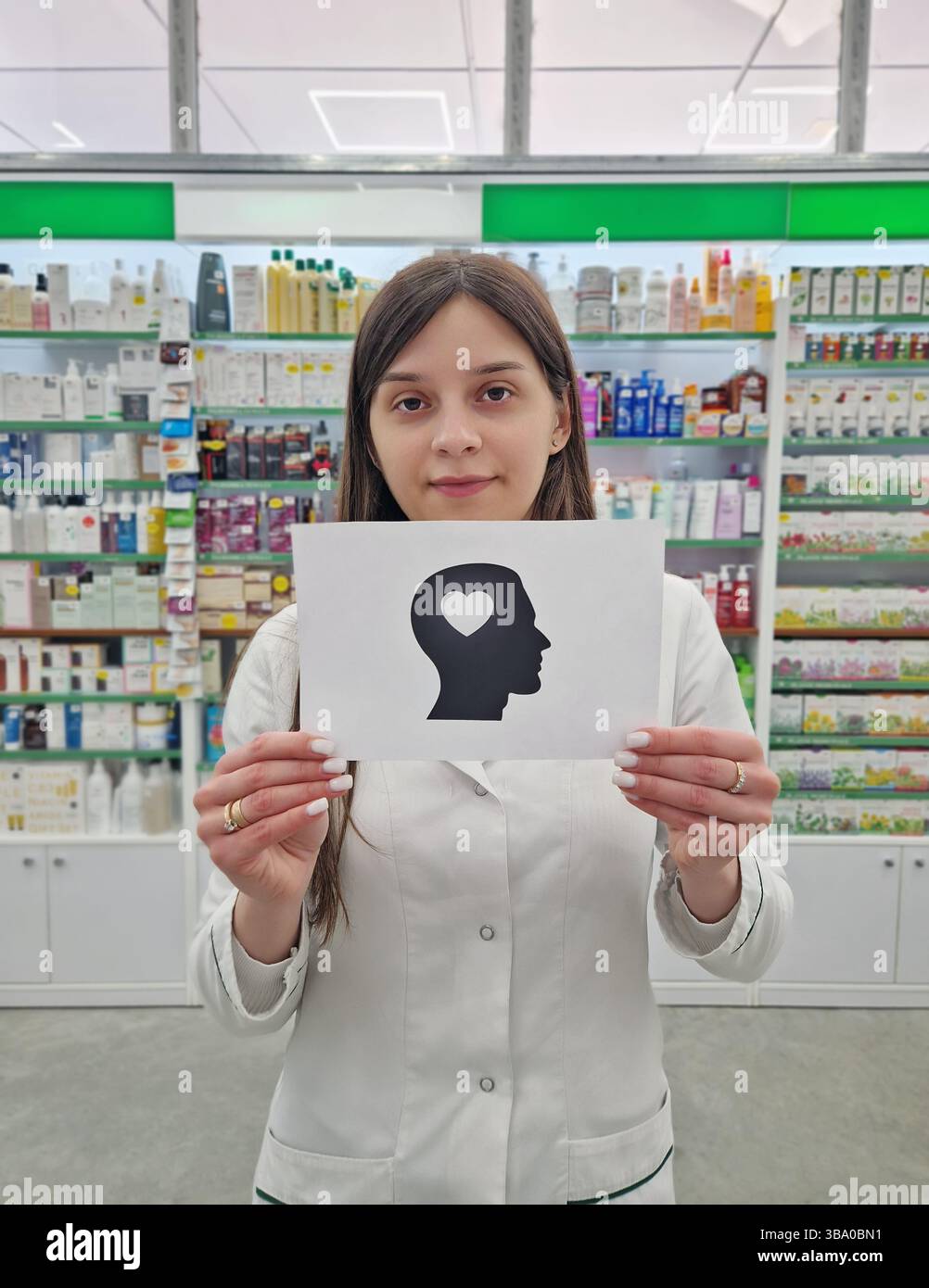 Farmacista donna che tiene in mano una carta con una silhouette della testa con un cuore all'interno, che simboleggia la salute mentale e il benessere, sopra gli scaffali della farmacia dietro Foto Stock