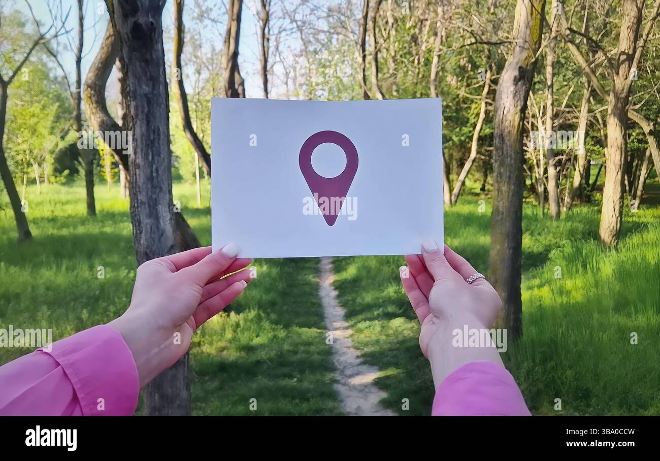 Mani femminili che tengono un foglio di carta bianco con il simbolo del perno di posizionamento su un percorso nella foresta. Guida di viaggio, scoperta ed esplorazioni a persona persa Foto Stock