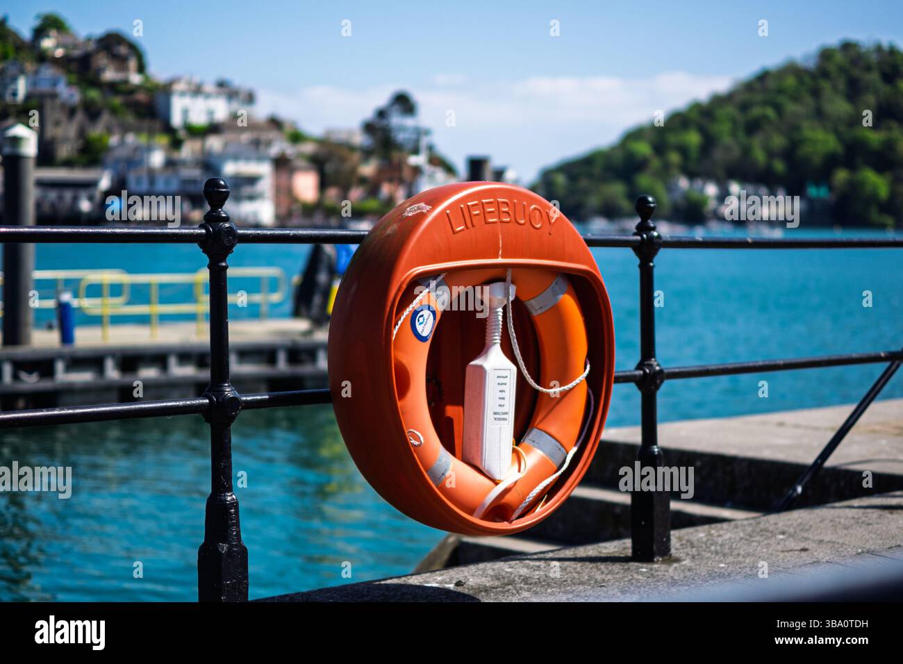 Un salvagente arancione luminoso è appeso su una ringhiera di ferro nera che si affaccia sull'acqua. Il salvagente è dotato di una fune e di un dispositivo di flottazione Foto Stock