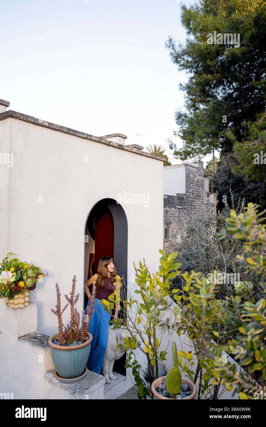 Una donna e il suo cane bianco escono su un giardino sul tetto circondato da piante in vaso e architettura in pietra, evocando un tranquillo stile di vita mediterraneo e un legame con la natura Foto Stock