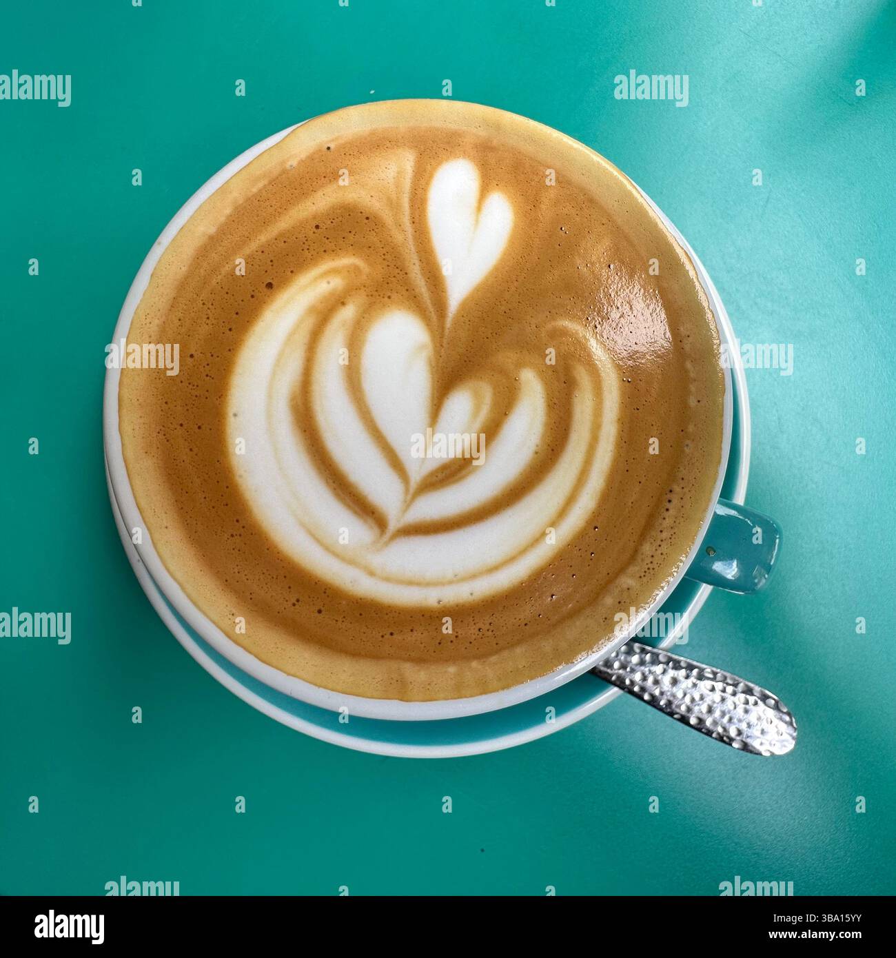 Caffè bianco piatto con cuore latte art servito in una tazza di tè su un tavolo turchese. Brunch luminoso in stile minimalista. Foto Stock