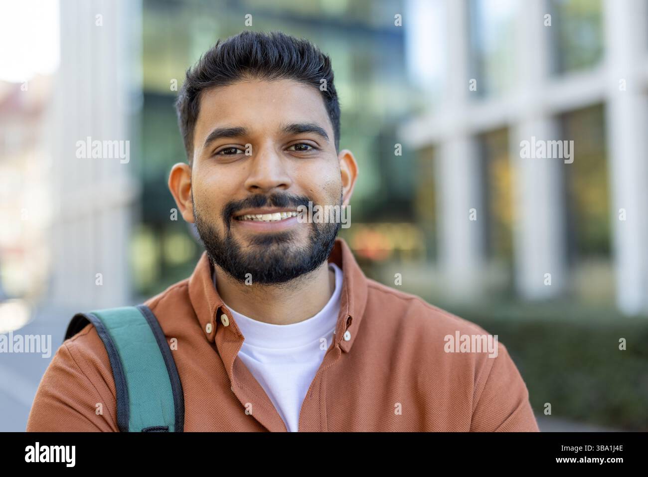 Un uomo indiano sorridente pone con sicurezza di fronte a un edificio moderno, possibilmente uno studente o un professionista con uno zaino. Foto Stock