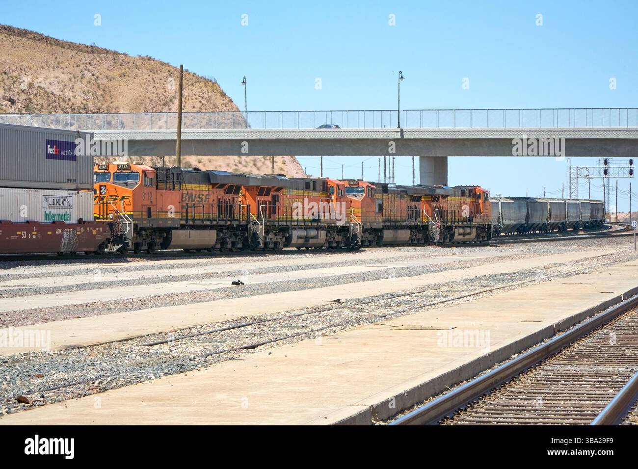 7 maggio 2025, Barstow, California, Stati Uniti: Fotografata nel cantiere ferroviario di Barstow, questa linea di locomotive della BNSF Railway si trova al di sotto di un cavalcavia del deserto, arancione brillante e nero contro le sfumature del Mojave. Questi cavalli da lavoro General Electric fanno parte del ritmo quotidiano di uno dei corridoi merci più trafficati negli Stati uniti occidentali.attaccati a una lunga serie di tramogge coperte, che probabilmente trasportano grano o materiali secchi sfusi, le locomotive riflettono la portata e l'efficienza della moderna logistica ferroviaria. Barstow funge da nodo critico nel sistema BNSF, dove i treni sono ri-cr Foto Stock