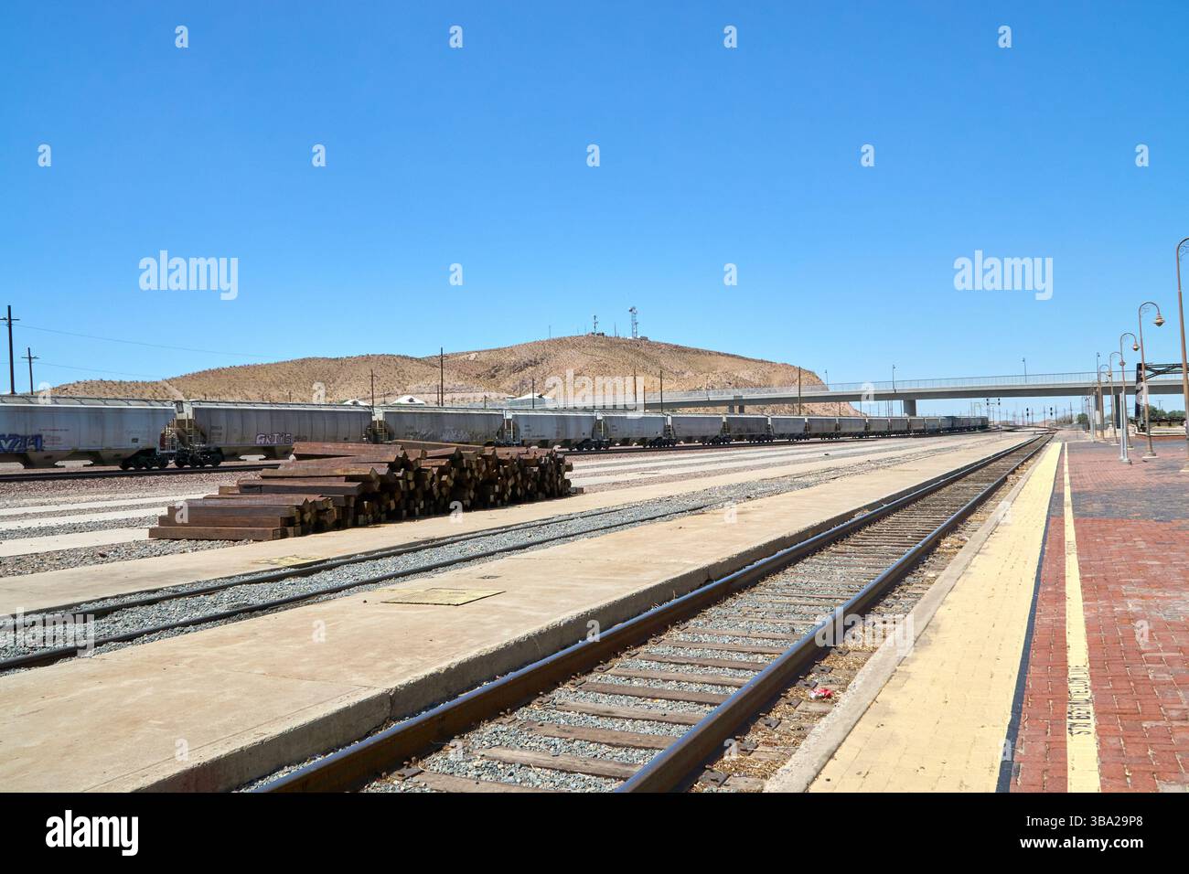 Barstow, California, Stati Uniti. 7 maggio 2025. Fotografata dal bordo della piattaforma Amtrak di Barstow, questa scena cattura l'ampia estensione di uno dei più grandi e trafficati cantieri ferroviari degli Stati uniti occidentali. Una serie di tramogge coperte si estende in lontananza sotto un cielo desertico, mentre pile di ferrovie inutilizzate attendono silenziosamente accanto ai binari.questa vista''”vuota di persone ma densa di infrastrutture''”rivela la portata e l'intensità silenziosa del ruolo di Barstow nel movimento merci americano. La piattaforma in mattoni rossi contrasta con le viuzze pallide e polverose di acciaio e cemento che si estendono Foto Stock
