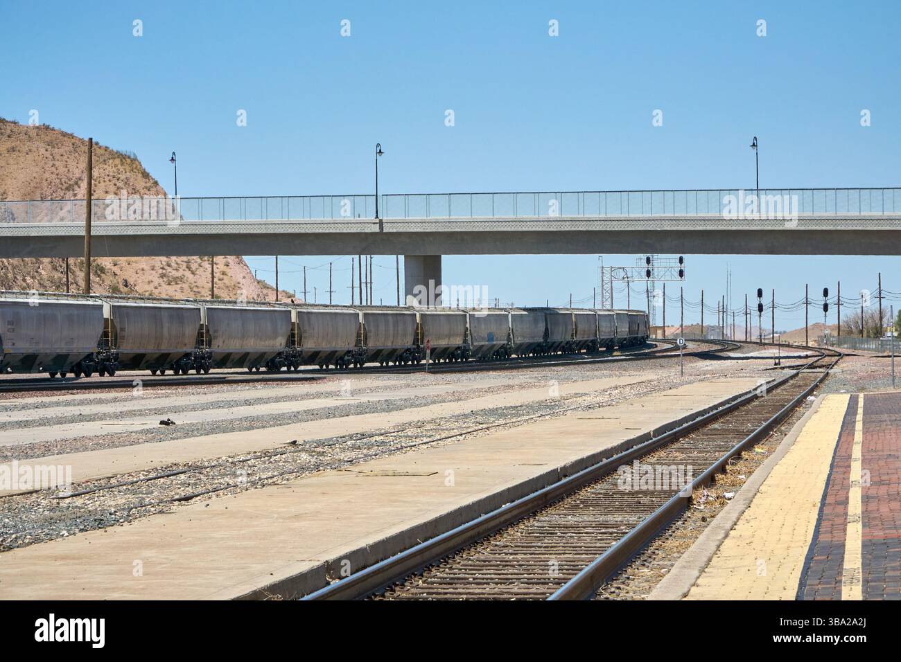 Barstow, California, Stati Uniti. 7 maggio 2025. Fotografata dal bordo della piattaforma Amtrak di Barstow, questa scena cattura l'ampia estensione di uno dei più grandi e trafficati cantieri ferroviari degli Stati uniti occidentali. Una serie di tramogge coperte si estende in lontananza sotto un cielo desertico, mentre pile di ferrovie inutilizzate attendono silenziosamente accanto ai binari.questa vista''”vuota di persone ma densa di infrastrutture''”rivela la portata e l'intensità silenziosa del ruolo di Barstow nel movimento merci americano. La piattaforma in mattoni rossi contrasta con le viuzze pallide e polverose di acciaio e cemento che si estendono Foto Stock