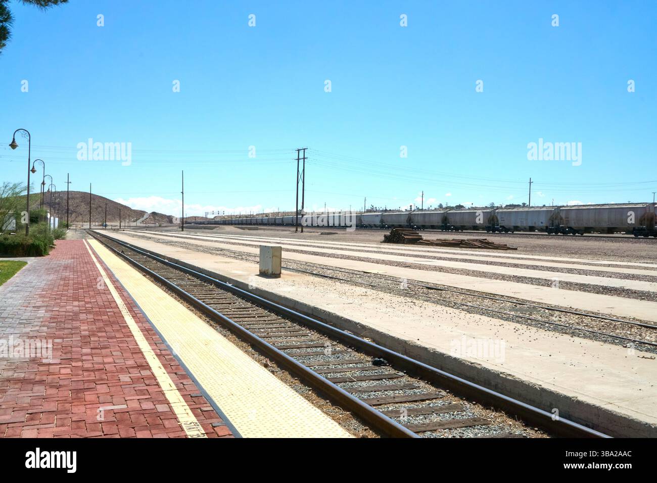 Barstow, California, Stati Uniti. 7 maggio 2025. Fotografata dal bordo della piattaforma Amtrak di Barstow, questa scena cattura l'ampia estensione di uno dei più grandi e trafficati cantieri ferroviari degli Stati uniti occidentali. Una serie di tramogge coperte si estende in lontananza sotto un cielo desertico, mentre pile di ferrovie inutilizzate attendono silenziosamente accanto ai binari.questa vista''”vuota di persone ma densa di infrastrutture''”rivela la portata e l'intensità silenziosa del ruolo di Barstow nel movimento merci americano. La piattaforma in mattoni rossi contrasta con le viuzze pallide e polverose di acciaio e cemento che si estendono Foto Stock