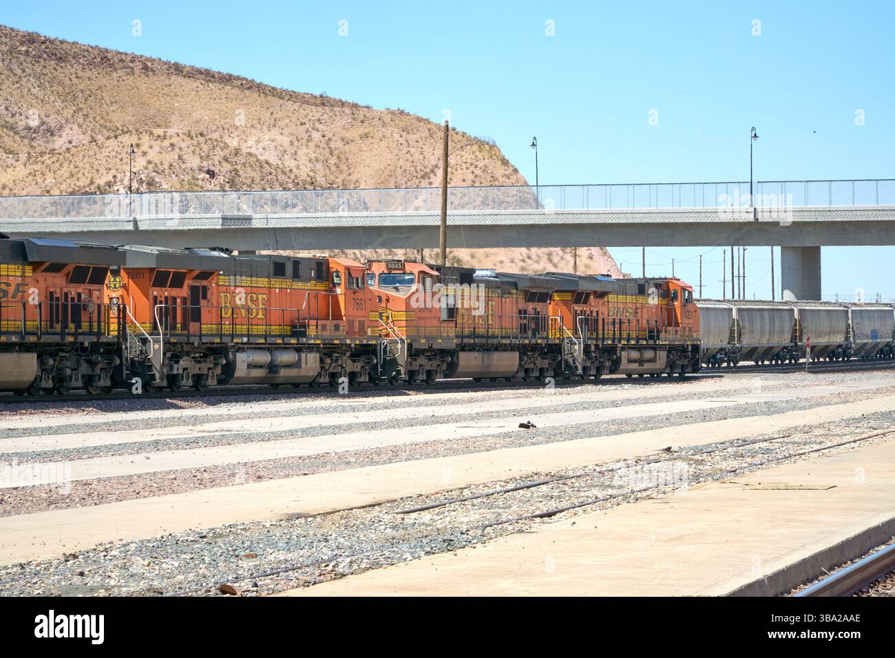 7 maggio 2025, Barstow, California, Stati Uniti: Fotografata nel cantiere ferroviario di Barstow, questa linea di locomotive della BNSF Railway si trova al di sotto di un cavalcavia del deserto, arancione brillante e nero contro le sfumature del Mojave. Questi cavalli da lavoro General Electric fanno parte del ritmo quotidiano di uno dei corridoi merci più trafficati negli Stati uniti occidentali.attaccati a una lunga serie di tramogge coperte, che probabilmente trasportano grano o materiali secchi sfusi, le locomotive riflettono la portata e l'efficienza della moderna logistica ferroviaria. Barstow funge da nodo critico nel sistema BNSF, dove i treni sono ri-cr Foto Stock
