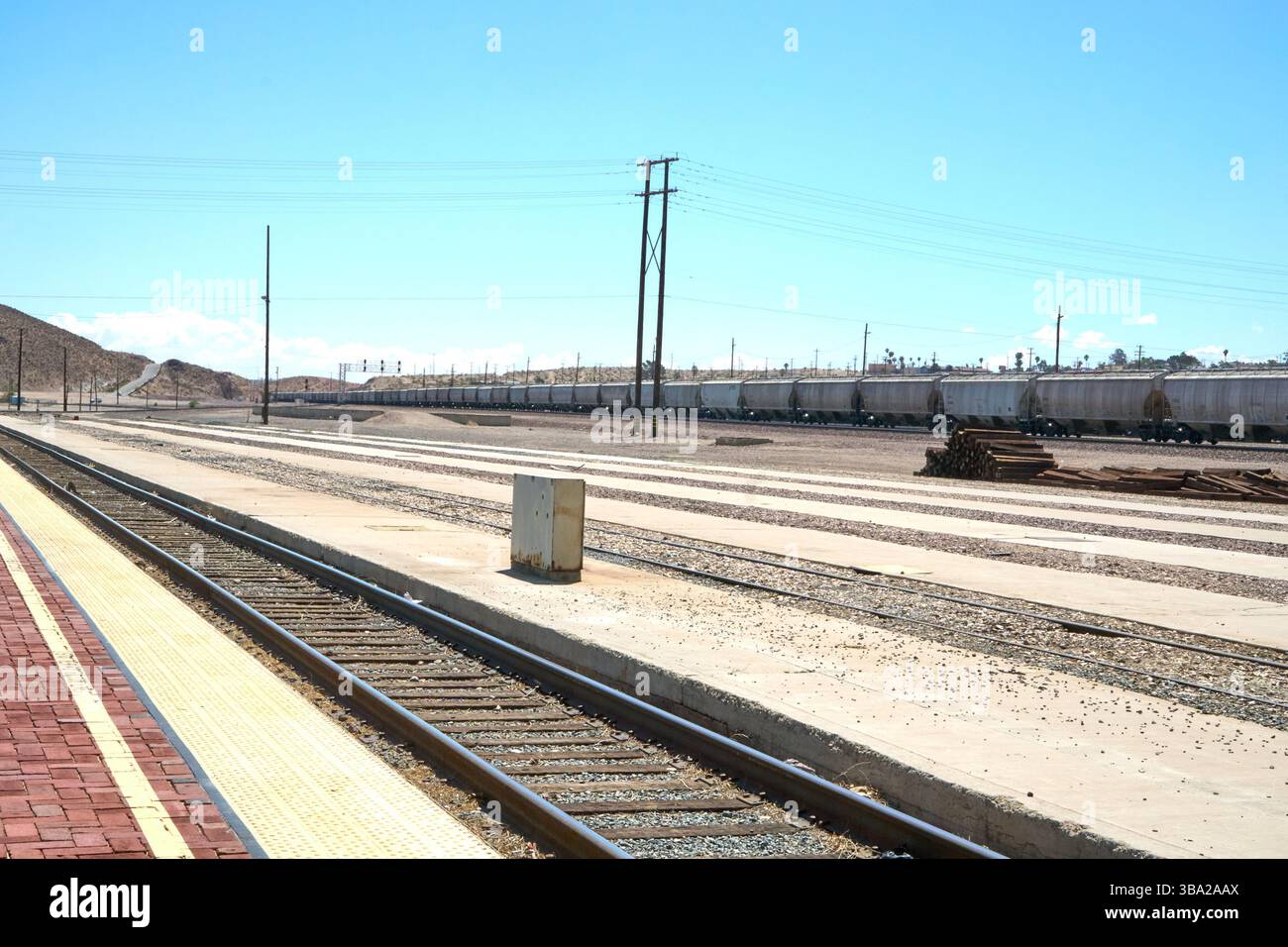 Barstow, California, Stati Uniti. 7 maggio 2025. Fotografata dal bordo della piattaforma Amtrak di Barstow, questa scena cattura l'ampia estensione di uno dei più grandi e trafficati cantieri ferroviari degli Stati uniti occidentali. Una serie di tramogge coperte si estende in lontananza sotto un cielo desertico, mentre pile di ferrovie inutilizzate attendono silenziosamente accanto ai binari.questa vista''”vuota di persone ma densa di infrastrutture''”rivela la portata e l'intensità silenziosa del ruolo di Barstow nel movimento merci americano. La piattaforma in mattoni rossi contrasta con le viuzze pallide e polverose di acciaio e cemento che si estendono Foto Stock