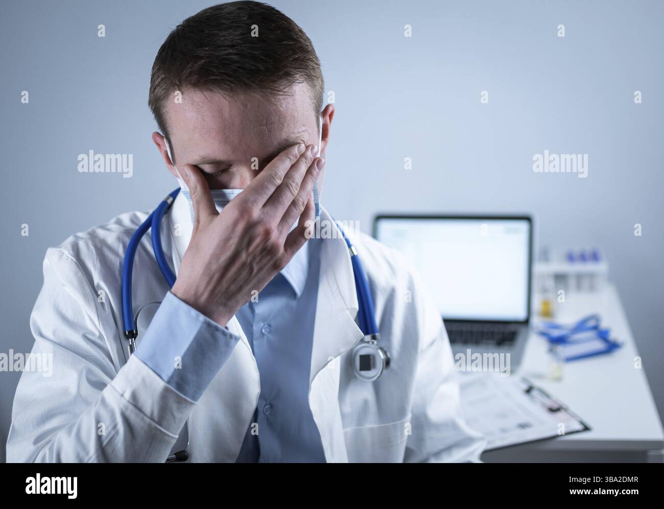 Esausto giovane medico in laboratorio cappotto toglie maschera e occhiali, sfrega gli occhi, ha mal di testa da affaticamento e overexertion dal lavoro in turno pesante in ho Foto Stock