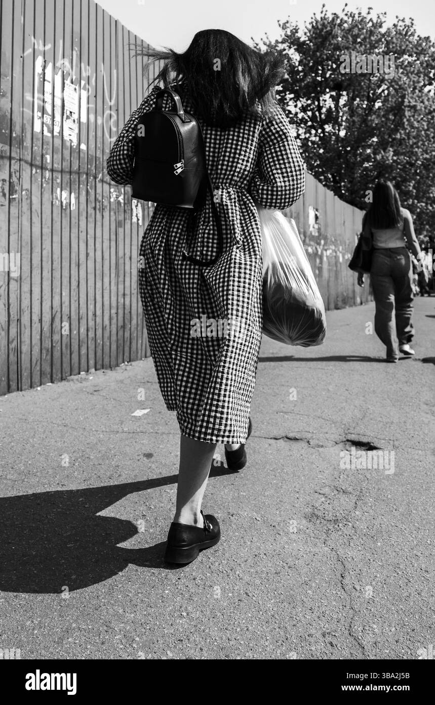Kiev, Ucraina - 1 maggio 2025: Foto in bianco e nero di una donna che si allontana dalla macchina fotografica di Kiev, portando con sé una borsa di plastica e uno zaino, durante una giornata di sole Foto Stock