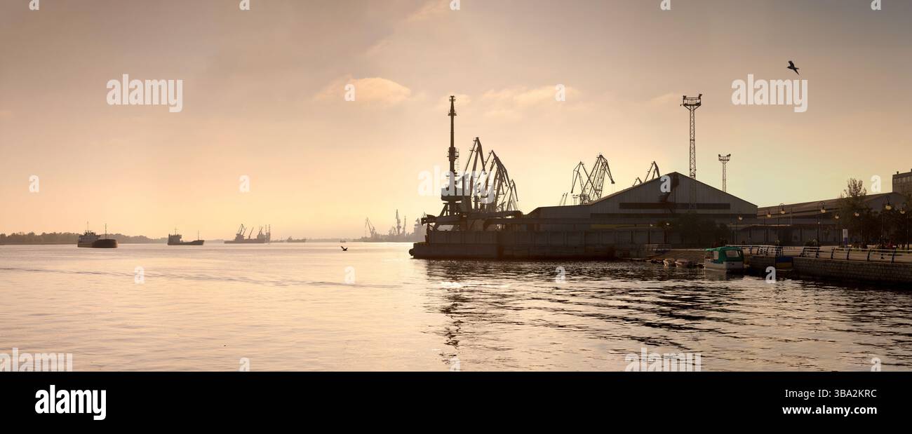 Il lungomare del porto marittimo nel centro di Kherson, Ucraina, con torreggianti gru portuali, navi ancorate e una passeggiata urbana. Ingresso porta industriale Foto Stock