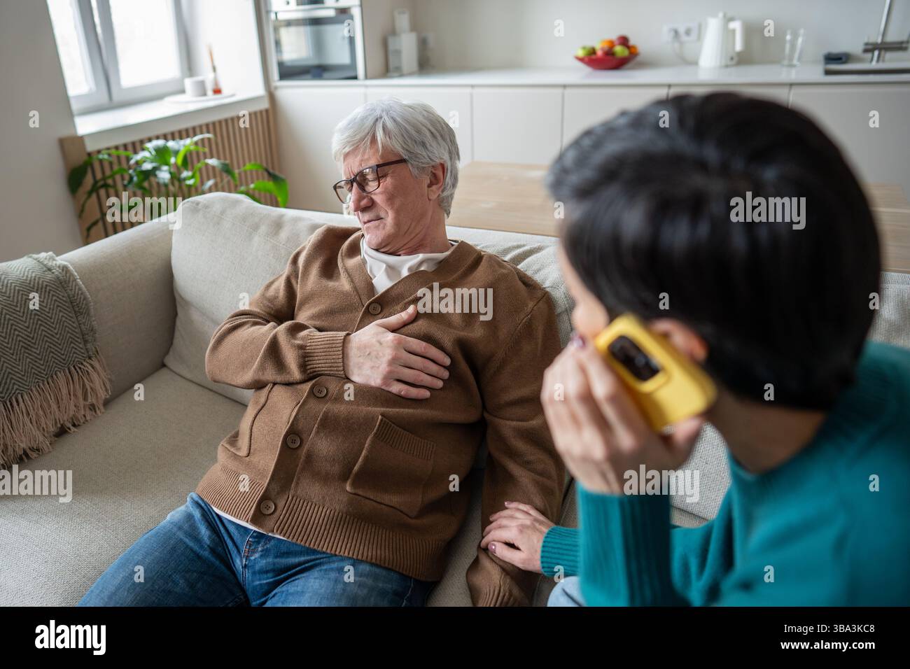 Uomo anziano che stringe il petto seduto sulla sedia, moglie chiama i servizi di emergenza con il telefono nelle vicinanze Foto Stock