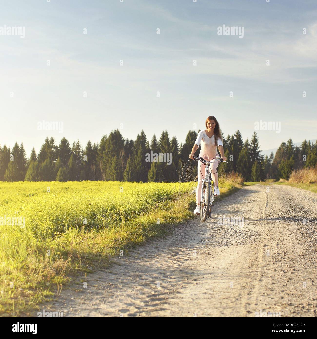 La giovane donna corre in bicicletta su una strada di campagna polverosa, le splende il sole del pomeriggio, vista frontale, VavriTovo, Slovacchia, Europa Foto Stock
