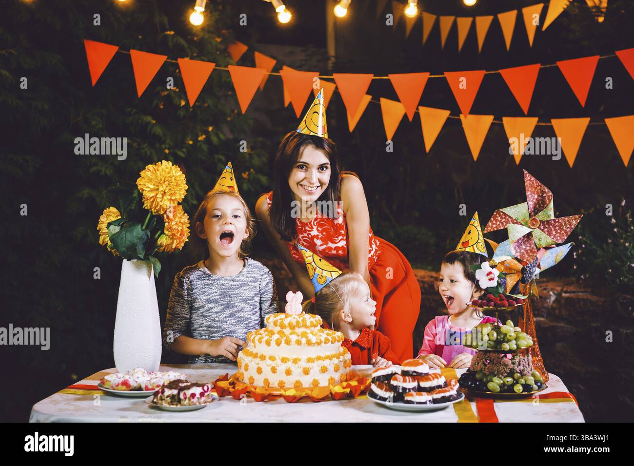 Una madre grande e tre bambini ad un tavolo festivo con dolci e una torta nel cortile decorato con luci e una ghirlanda di lampade. Il concetto Foto Stock
