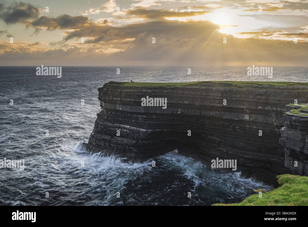 Pescatore in piedi e pesca dal bordo delle alte scogliere di Downpatrick Head con onde che si infrangono sotto la drammatica alba, contea di Mayo, Irlanda, UE Foto Stock