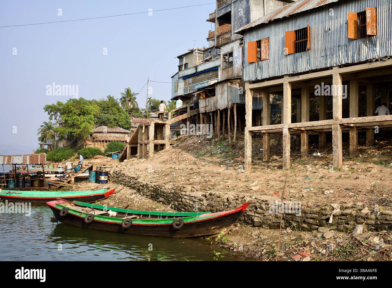 Palafitte su pilastri di cemento e legno fiancheggiano le rive del lago Kaptai a Rangamati, Bangladesh, affacciate su barche ormeggiate e detriti sparsi. Foto Stock
