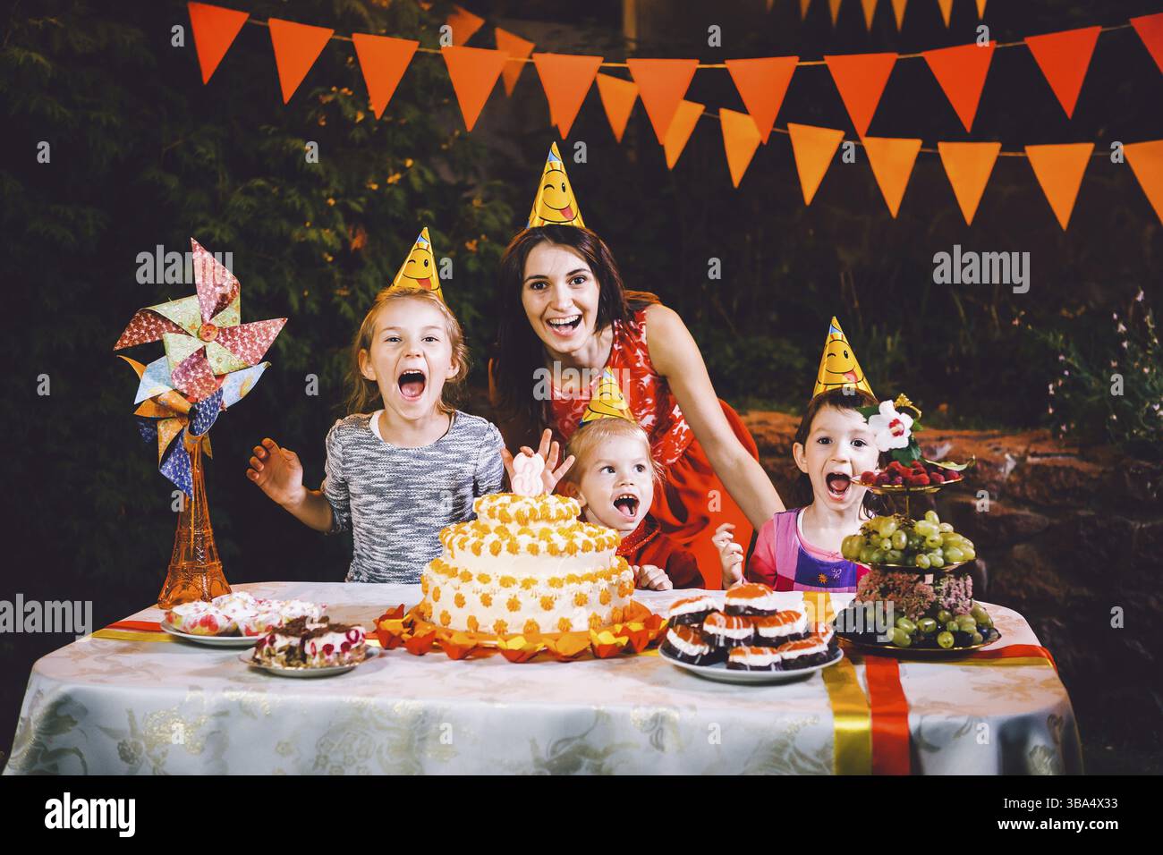 Una madre grande e tre bambini ad un tavolo festivo con dolci e una torta nel cortile decorato con luci e una ghirlanda di lampade. Il concetto Foto Stock