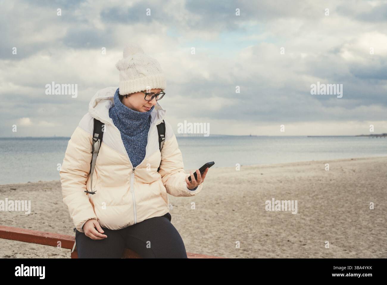 Donna caucasica in cappello e giacca con zaino in inverno siede su un molo di legno sulla spiaggia vicino al Mare del Nord. Danimarca Copenhagen turista usa un m Foto Stock