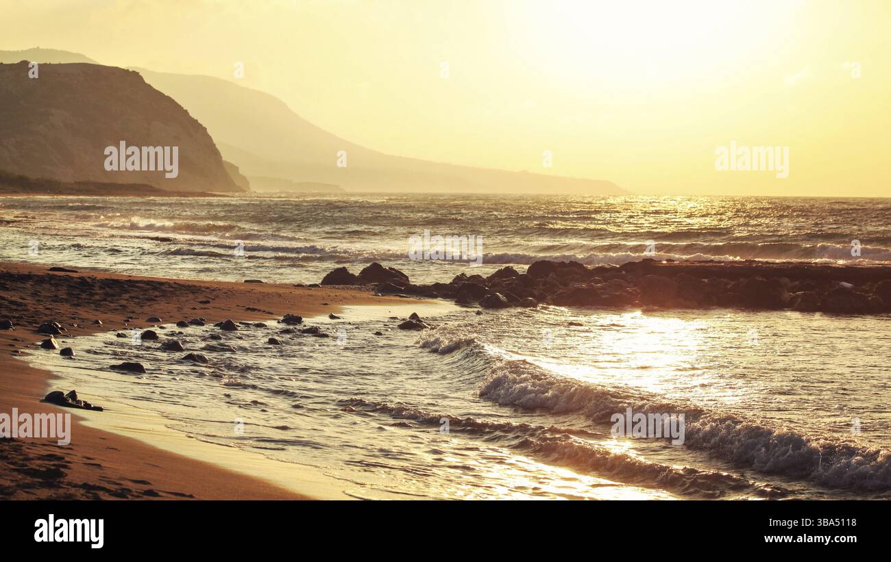 Spiaggia selvaggia vuota illuminata da un tramonto dorato. Piccole onde sulla roccia, colline sullo sfondo nebbioso, Lapta, Cipro, Europa Foto Stock