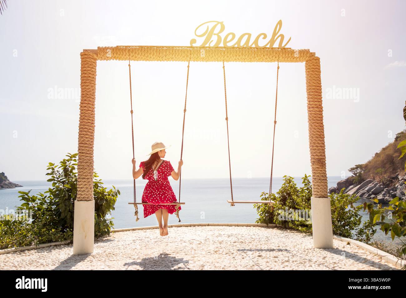 Vista posteriore della donna asiatica in maxi abito rosso seduta su un'altalena con un cartello "Beach" in cima, vista mare sullo sfondo. buon viaggio estivo Foto Stock