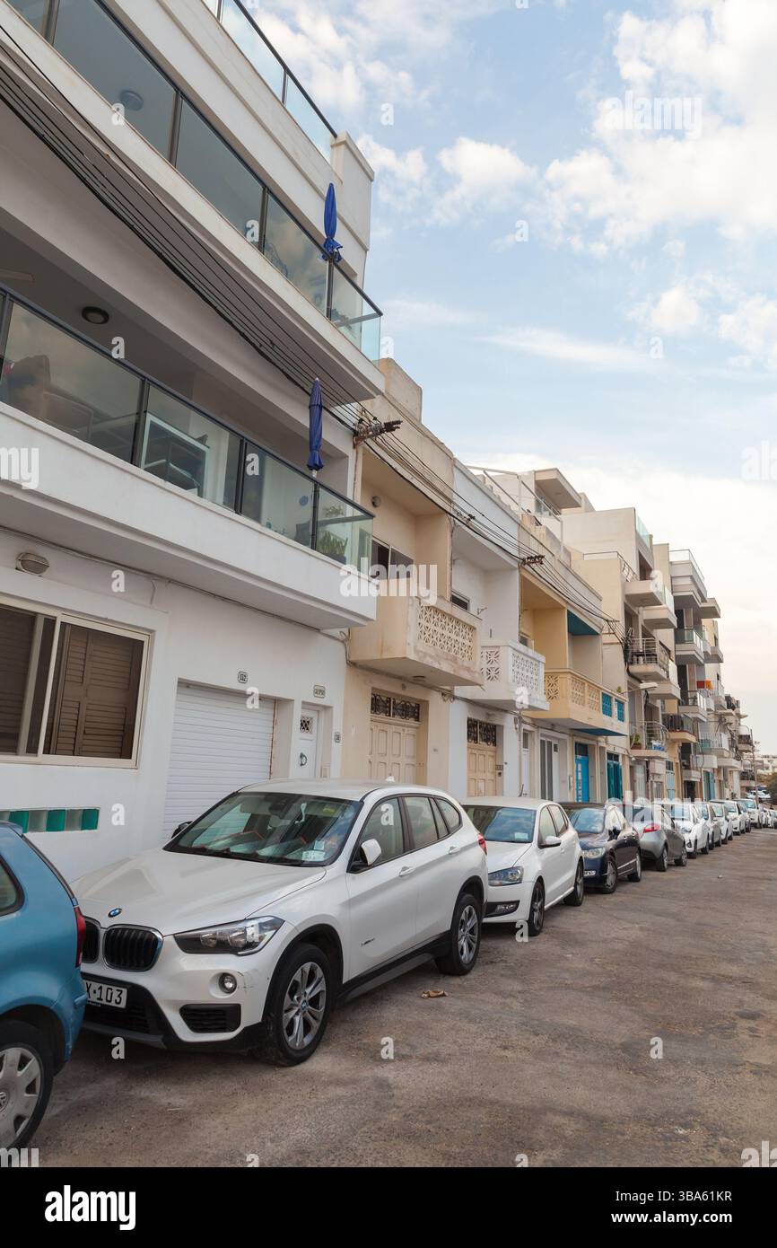 Mellieha, Malta - 29 agosto 2019: Vista verticale su strada di una strada urbana fiancheggiata da appartamenti residenziali e auto parcheggiate sotto la luce del giorno Foto Stock