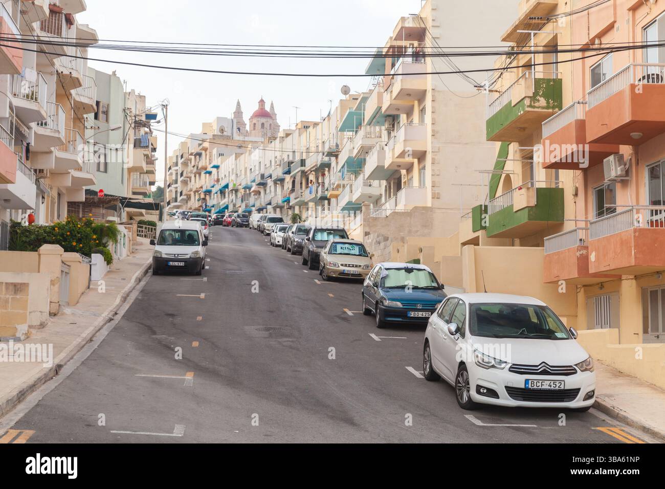 Mellieha, Malta - 29 agosto 2019: Vista di una strada urbana in pendenza fiancheggiata da colorati appartamenti residenziali e auto parcheggiate sotto una amb Foto Stock